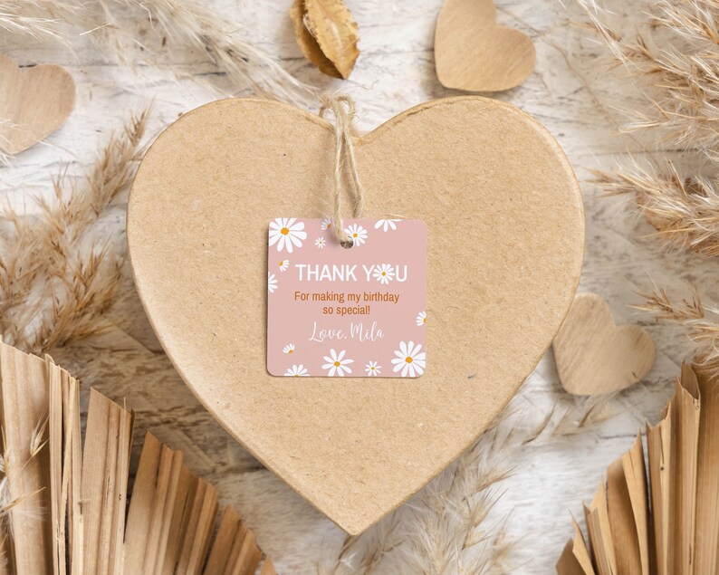 May include: A square pink gift tag with white daisy illustrations and the text "THANK YOU For making my birthday so special! Love, Mila" printed in white and orange. The tag is attached with a jute string to a large, heart-shaped cardboard box.