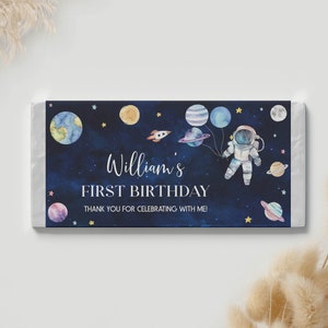 May include: A blue and white chocolate bar wrapper with a watercolor illustration of an astronaut holding balloons shaped like planets. The text on the wrapper reads "William's First Birthday" and "Thank you for celebrating with me!"