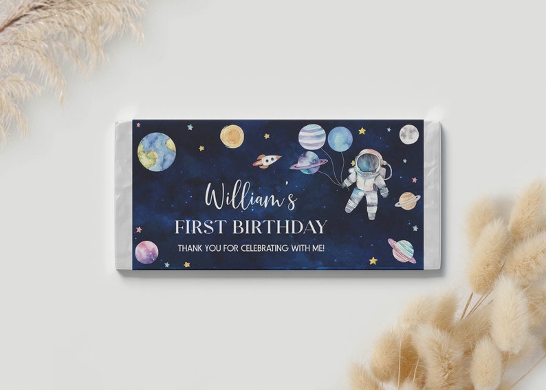 May include: A blue and white chocolate bar wrapper with a watercolor illustration of an astronaut holding balloons shaped like planets. The text on the wrapper reads "William's First Birthday" and "Thank you for celebrating with me!"