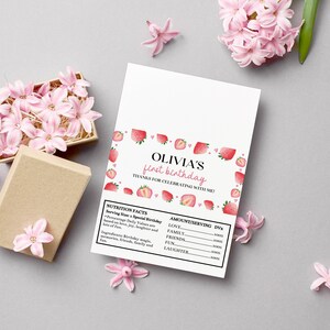 May include: A rectangular white card featuring a strawberry-themed border and the text OLIVIA'S first birthday THANKS FOR CELEBRATING WITH ME! Below is a nutrition facts label listing love, family, friends, fun, and laughter as ingredients.