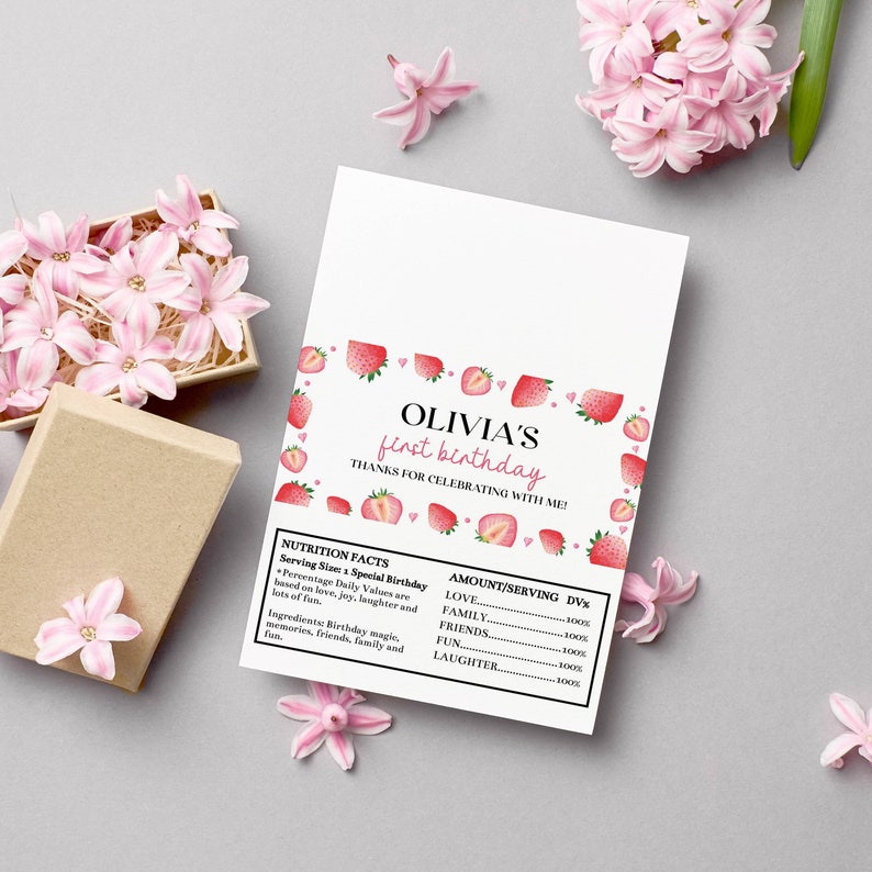 May include: A rectangular white card featuring a strawberry-themed border and the text OLIVIA'S first birthday THANKS FOR CELEBRATING WITH ME! Below is a nutrition facts label listing love, family, friends, fun, and laughter as ingredients.
