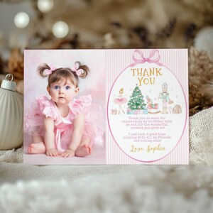May include: A rectangular thank you card featuring a photograph of a toddler in a pink ruffled dress on the left and a pink striped background with a gold oval frame on the right. The frame contains a Christmas tree, a nutcracker, and text.