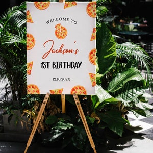 May include: A white sign with a pizza theme welcomes guests to Jackson's 1st Birthday party on 12.10.202X. The sign is on a wooden easel.