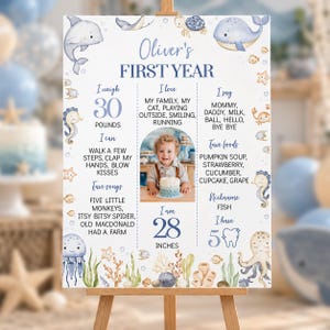 May include: A white milestone birthday sign titled Oliver's First Year, featuring blue watercolor sea creatures like whales, dolphins, and octopuses. It includes text sections for weight, height, favorite foods, songs, and a central photo of a child.
