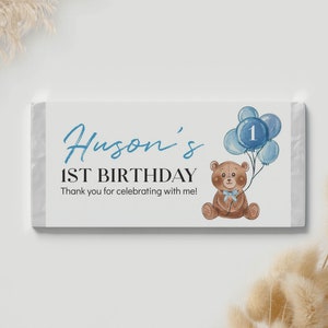 May include: A rectangular candy bar wrapper featuring a white background with the blue text Huson's 1ST BIRTHDAY and Thank you for celebrating with me! A brown teddy bear with a light blue bow holds a cluster of blue balloons with the number 1.