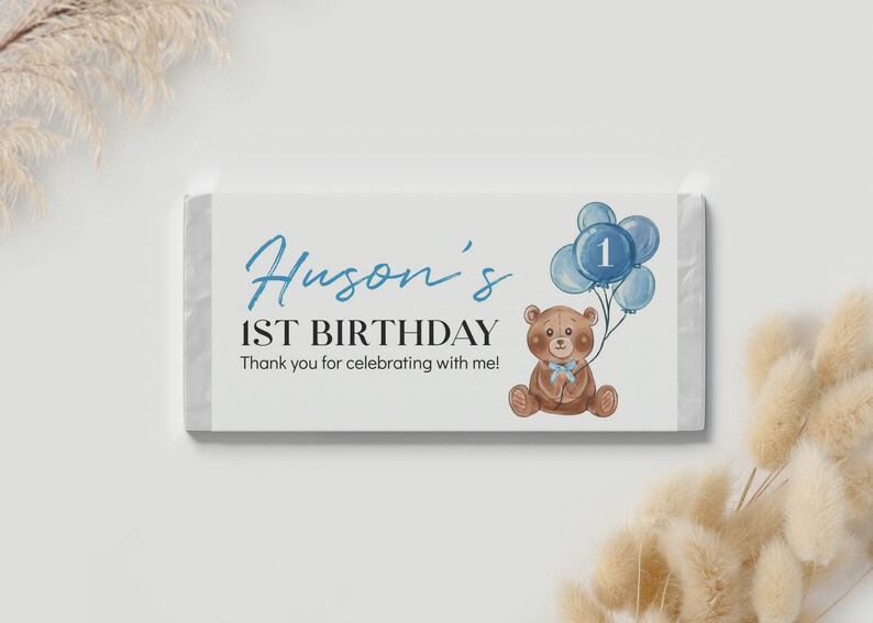 May include: A rectangular candy bar wrapper featuring a white background with the blue text Huson's 1ST BIRTHDAY and Thank you for celebrating with me! A brown teddy bear with a light blue bow holds a cluster of blue balloons with the number 1.