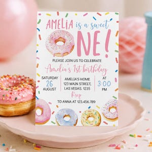 May include: A rectangular birthday invitation card featuring a pink donut illustration, colorful sprinkles, and text that reads: AMELIA is a sweet ONE! PLEASE JOIN US TO CELEBRATE Amelia's 1st birthday SATURDAY 26 AUGUST AT 3:00 PM.