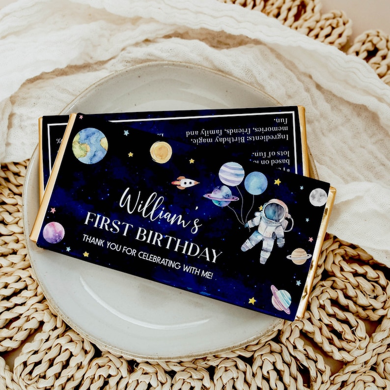 May include: A blue and black chocolate bar with a space theme. It features an astronaut, planets, stars, and the text "William's First Birthday. Thank you for celebrating with me!"