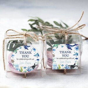 Pu&ograve; includere: Due scatoline quadrate trasparenti legate con spago marrone, contenenti ciascuna un macaron viola chiaro e fogliame verde. Ogni scatola presenta un'etichetta con animali marini ad acquerello e il testo THANK YOU for celebrating with us! Love, Huson.
