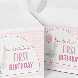 May include: Two white cardboard gable boxes featuring a pink gingham pattern and a central arch graphic. The design includes a watercolor illustration of a goose wearing a party hat, holding a pink balloon with the number one, and the text Amelia's FIRST BIRTHDAY.