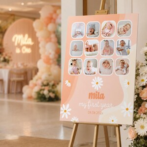 May include: A peach-colored sign on a wooden easel, featuring a collage of baby photos. The sign has a daisy and wave design, with the text "mila my first year" and the date 09.08.202x. The background includes party decorations.