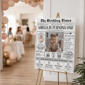 May include: A white rectangular birthday milestone sign titled The Birthday Times featuring a central photograph of a baby, text detailing birth date, height of 66 cm, weight of 13.6 kg, favourite foods, songs, books, and toys in black ink.