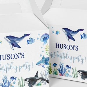 May include: Two white cardboard favor boxes featuring watercolor illustrations of sea creatures including a whale, shark, orca, turtle, jellyfish, and fish. The text HUSON'S first birthday party! is printed in blue on the front of each box.