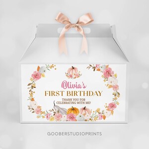 May include: A white gable box with a satin ribbon bow on top. The front features a floral wreath design with pumpkins and the text: Olivia's FIRST BIRTHDAY THANK YOU FOR CELEBRATING WITH ME! The bottom includes the text GOOBERSTUDIOPRINTS.