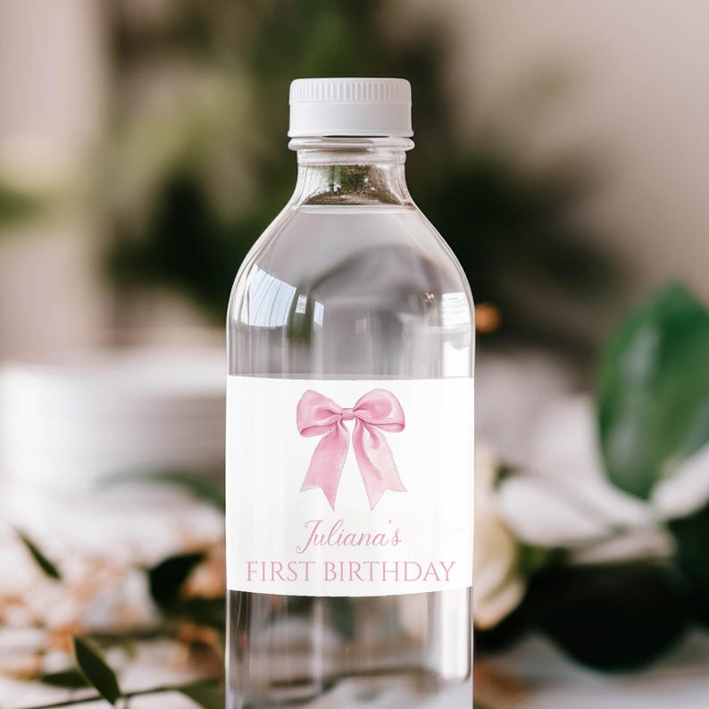 Puede incluir: Una botella de agua de pl&aacute;stico transparente con tapa blanca y una etiqueta rectangular. La etiqueta presenta una ilustraci&oacute;n de un lazo rosa en acuarela sobre el texto Juliana's FIRST BIRTHDAY en una fuente cursiva rosa y una fuente sans-serif.