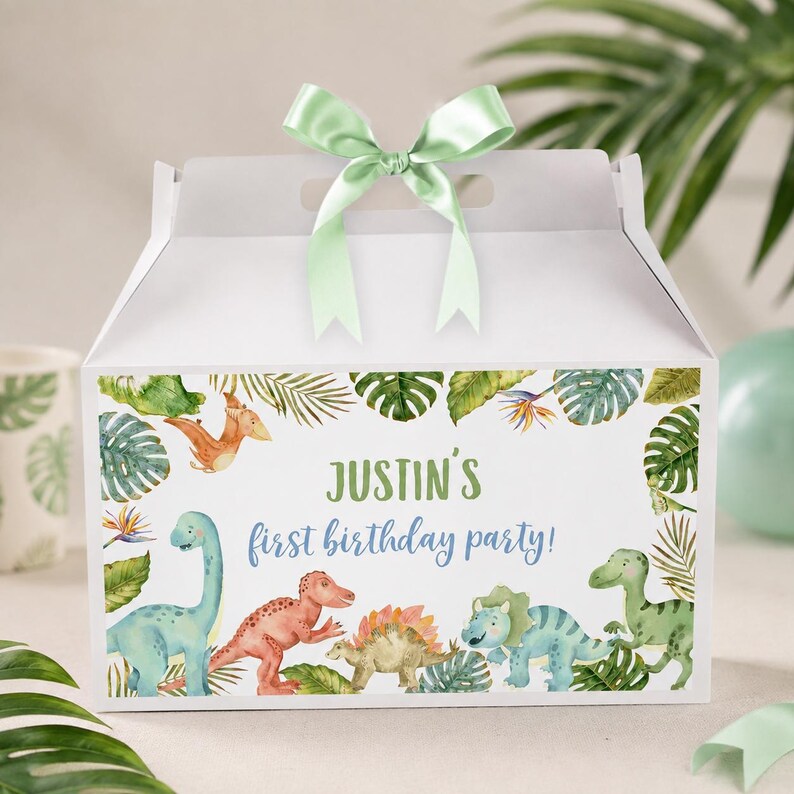 May include: A white gable box with a light green satin ribbon bow on top. The front features a watercolor illustration of various dinosaurs and tropical leaves with the text "JUSTIN'S first birthday party!" in green and blue script lettering.