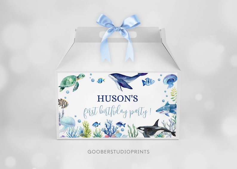 May include: A white gable box with a light blue satin ribbon on top, featuring a watercolor ocean theme with a sea turtle, whale, shark, orca, jellyfish, and various fish. The text reads HUSON'S first birthday party! in blue lettering.