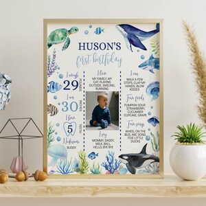 May include: A blue and white first birthday milestone chart with an underwater theme. The chart includes a photo of a baby, a list of milestones, and a list of favorite foods and songs. The chart is decorated with sea creatures, including a whale, a turtle, a shark, and a jellyfish.