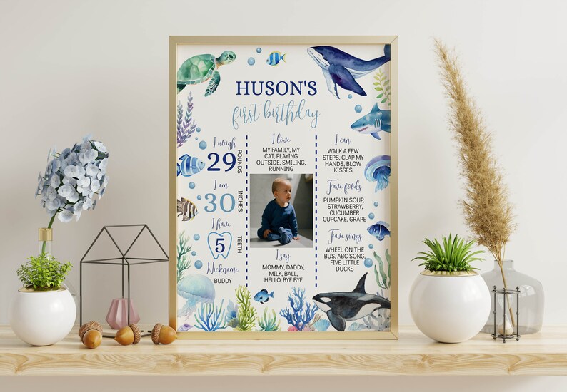May include: A blue and white first birthday milestone chart with an underwater theme. The chart includes a photo of a baby, a list of milestones, and a list of favorite foods and songs. The chart is decorated with sea creatures, including a whale, a turtle, a shark, and a jellyfish.