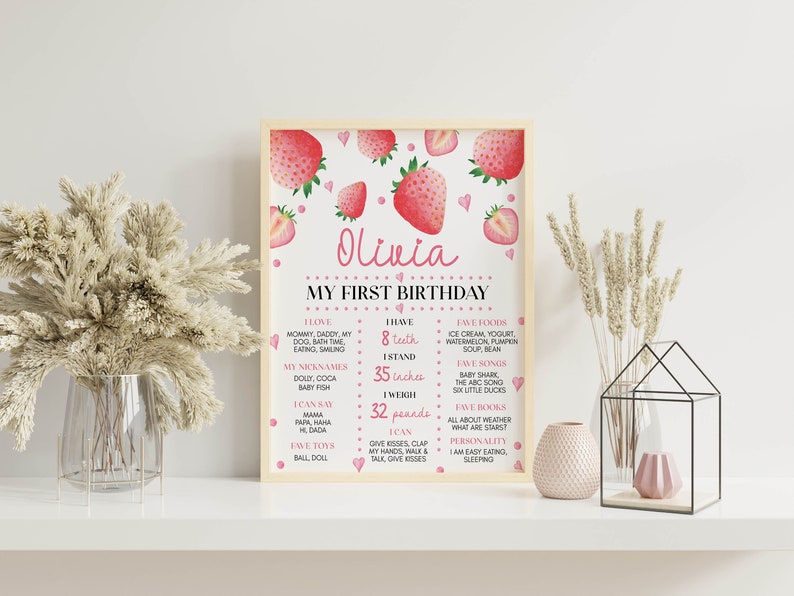 May include: A framed personalized first birthday milestone sign featuring a strawberry theme with pink and red illustrations. The text includes the name Olivia, My First Birthday, and lists developmental milestones with 8 teeth, 35 inches, and 32 pounds.