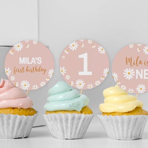 May include: Three cupcakes in white paper liners topped with pink, light blue, and yellow frosting. Behind each cupcake is a round pink topper featuring white daisy illustrations and text reading Mila's first birthday, 1, and Mila is ONE.