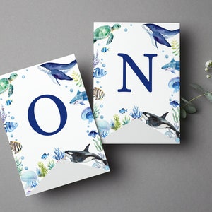 May include: Two white pennant flags featuring watercolor illustrations of sea creatures including a whale, sea turtle, shark, orca, and jellyfish. The left flag displays a large dark blue letter O, and the right flag displays a large dark blue letter N.