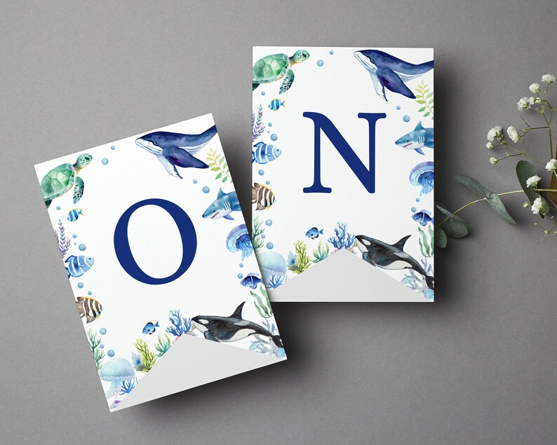 May include: Two white pennant flags featuring watercolor illustrations of sea creatures including a whale, sea turtle, shark, orca, and jellyfish. The left flag displays a large dark blue letter O, and the right flag displays a large dark blue letter N.