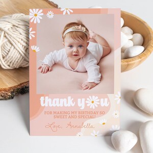 May include: A rectangular thank you card featuring a photo of a baby wearing a white headband, surrounded by a peach-colored border with white daisy illustrations. The text reads: thank you FOR MAKING MY BIRTHDAY SO SWEET AND SPECIAL! Love, Annabella.