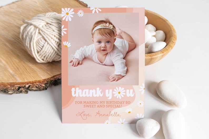 May include: A rectangular thank you card featuring a photo of a baby wearing a white headband, surrounded by a peach-colored border with white daisy illustrations. The text reads: thank you FOR MAKING MY BIRTHDAY SO SWEET AND SPECIAL! Love, Annabella.