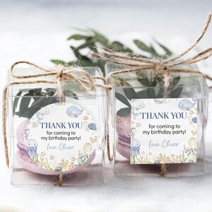 May include: Two clear plastic favor boxes tied with twine, each containing a pink macaron and greenery. Each box has a circular or square tag with the text "THANK YOU for coming to my birthday party! Love, Oliver" and sea creature illustrations.