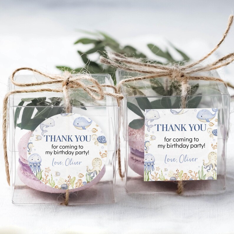 May include: Two clear plastic favor boxes tied with twine, each containing a pink macaron and greenery. Each box has a circular or square tag with the text "THANK YOU for coming to my birthday party! Love, Oliver" and sea creature illustrations.