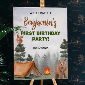 Op de afbeelding: Een rechthoekig welkomstbord voor een eerste verjaardagsfeest met een bosthema, inclusief een bruine tent, kampvuur, eekhoorn en beer. De tekst luidt: WELCOME TO Benjamin's FIRST BIRTHDAY PARTY! 20.10.202X. De achtergrond toont diverse dennenbomen.