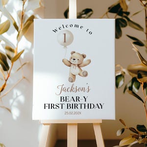 May include: A white rectangular sign featuring a brown teddy bear holding a beige balloon with the number 1. The text reads Welcome to, Jackson's, BEAR-Y FIRST BIRTHDAY, and 25.02.202X. The sign is displayed on a light wooden easel stand.