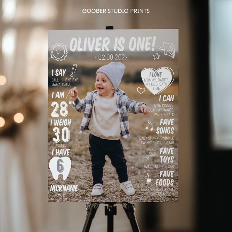 May include: A milestone birthday sign titled OLIVER IS ONE! featuring a photograph of a child in a grey beanie, plaid shirt, and dark pants. The sign includes text details like I SAY, I AM 28", I WEIGH 30 POUNDS, I HAVE 6 TEETH, I LOVE, I CAN, FAVE SONGS, FAVE TOYS, and FAVE FOODS.