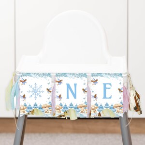 May include: A high chair banner featuring a winter theme with light blue snowflakes, small fairies, and mushroom illustrations. The banner displays the letters O, N, and E in a light blue font, accented with pastel-colored fabric tassels on both sides.