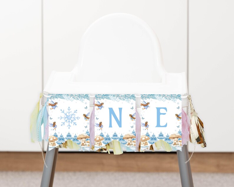 May include: A high chair banner featuring a winter theme with light blue snowflakes, small fairies, and mushroom illustrations. The banner displays the letters O, N, and E in a light blue font, accented with pastel-colored fabric tassels on both sides.
