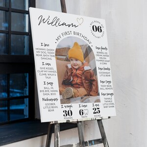 May include: A white sign with a photo of a baby boy in a red hat and brown jacket. The sign says "William, My First Birthday" and lists the baby's favorite foods, books, toys, and songs. It also includes his weight, 30 pounds, number of teeth, 6, and height, 25 inches.