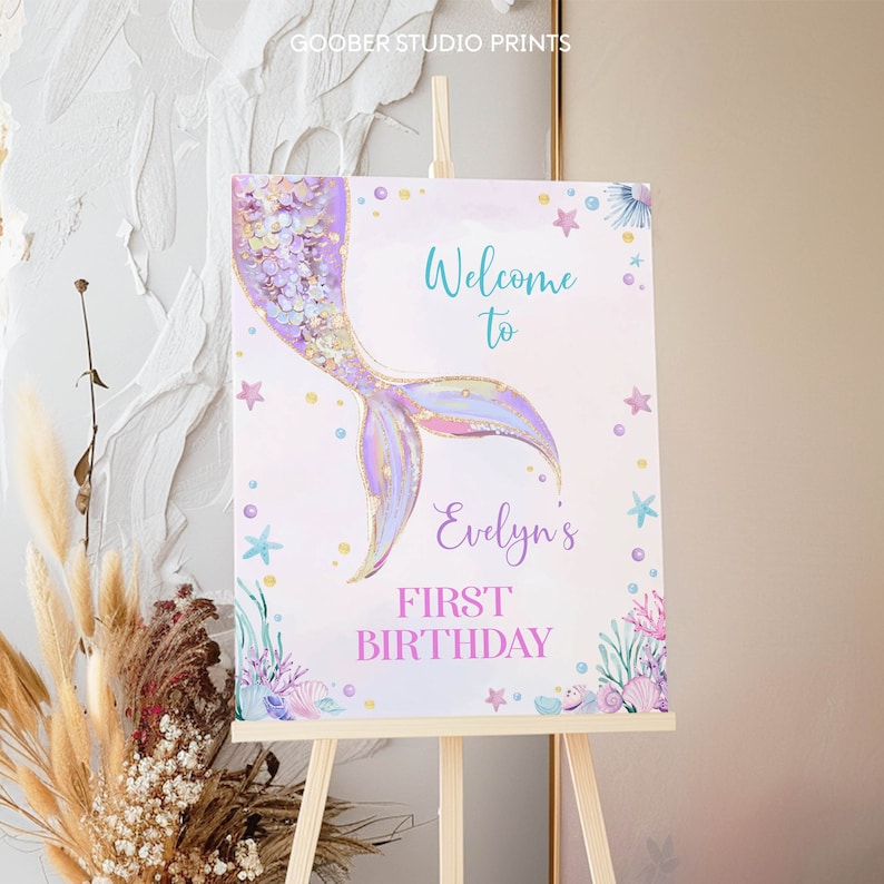 May include: A rectangular sign on a wooden easel featuring a watercolor mermaid tail in purple and gold. The text reads Welcome to Evelyn's FIRST BIRTHDAY in teal and purple. The design includes illustrations of starfish, shells, and coral.
