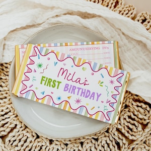 May include: A rectangular chocolate bar wrapper featuring a pastel striped background with a white scalloped center containing the text Mila's FIRST BIRTHDAY in pink and purple, surrounded by colorful confetti, hearts, and star shapes.