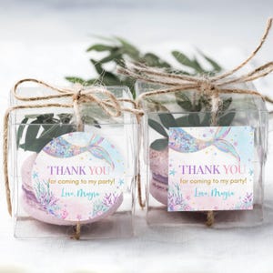 May include: Two clear favor boxes with a mermaid tail design and the text "Thank you for coming to my party! Love, Mayra" on each box. The boxes are tied with twine and contain a pink and white macaron.