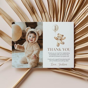 May include: A rectangular thank you card featuring a photo of a baby in a tan bear-ear onesie holding a beige balloon on the left. The right side shows a watercolor teddy bear with a balloon, the number 1, and text reading Thank You, Jackson.