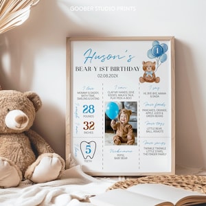 May include: A personalized first birthday milestone poster in a wooden frame featuring a brown teddy bear illustration, blue balloons, and text detailing baby facts including 28 pounds, 32 inches, 5 teeth, and a photo of a child in a bear suit.