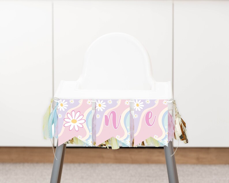 Op de afbeelding: Een kinderstoelbanner met drie wimpels op een golvende achtergrond in roze, lavendel en lichtblauw. De banner toont witte madeliefjes en de letters O, N en E in een roze schrijflettertype, geaccentueerd met hangende metallic gouden kwastjes.
