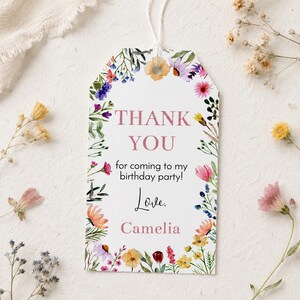 May include: A white rectangular gift tag with rounded top corners and a white string, featuring a watercolor floral border in shades of pink, yellow, blue, and green. The text reads: THANK YOU for coming to my birthday party! Love. Camelia.