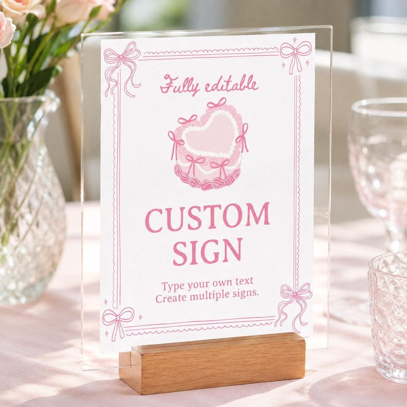 Peut inclure: Un porte-affiche en acrylique transparent avec une base en bois pr&eacute;sentant une carte blanche avec du texte et des graphismes roses. La carte comporte une illustration de g&acirc;teau en forme de c&oelig;ur, des n&oelig;uds roses et le texte Fully editable, CUSTOM SIGN.