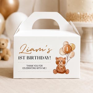 May include: A white cardboard gable box featuring a brown teddy bear illustration holding a cluster of balloons with the number one. The text reads Liam's 1ST BIRTHDAY! THANK YOU FOR CELEBRATING WITH ME! in brown and black lettering on the front.