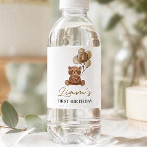 May include: A clear plastic water bottle with a white label featuring a brown teddy bear holding a cluster of balloons with the number 1 on the center balloon. The text Liam's FIRST BIRTHDAY is printed in gold and black script and block lettering.
