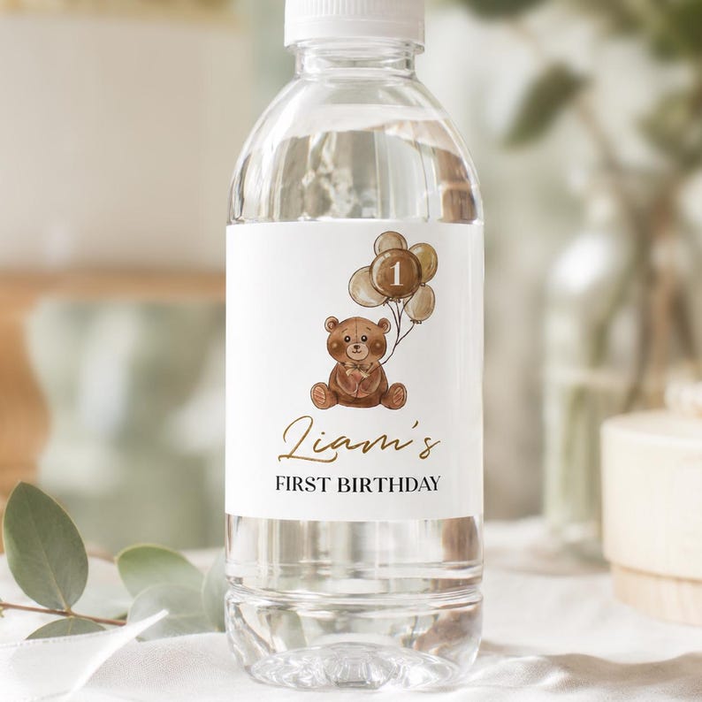 May include: A clear plastic water bottle with a white label featuring a brown teddy bear holding a cluster of balloons with the number 1 on the center balloon. The text Liam's FIRST BIRTHDAY is printed in gold and black script and block lettering.