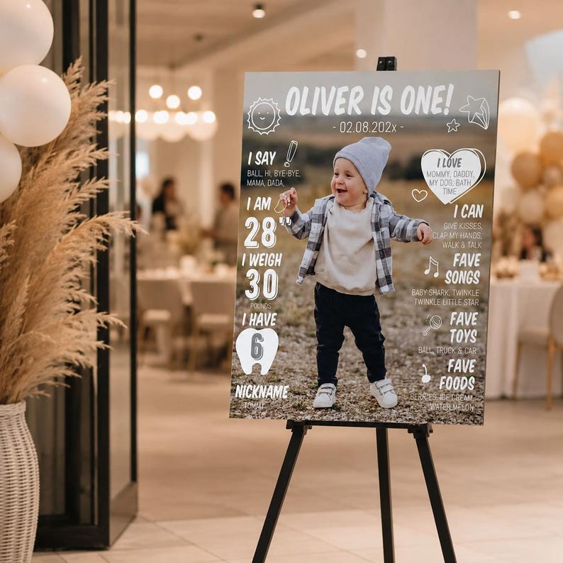 May include: A rectangular birthday milestone sign on a black easel featuring a photo of a toddler in a grey beanie and plaid shirt. The text reads OLIVER IS ONE, 02.08.202x, I SAY, I AM 28", I WEIGH 30 POUNDS, I HAVE 6 TEETH, I LOVE, I CAN, FAVE SONGS, FAVE TOYS, FAVE FOODS.