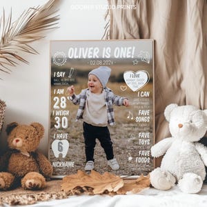 May include: A personalized milestone poster titled OLIVER IS ONE! featuring a central photograph of a toddler in a grey beanie, plaid shirt, and dark pants. The poster includes white text detailing stats like 28 inches, 30 pounds, 6 teeth, and favorite songs, toys, and foods.